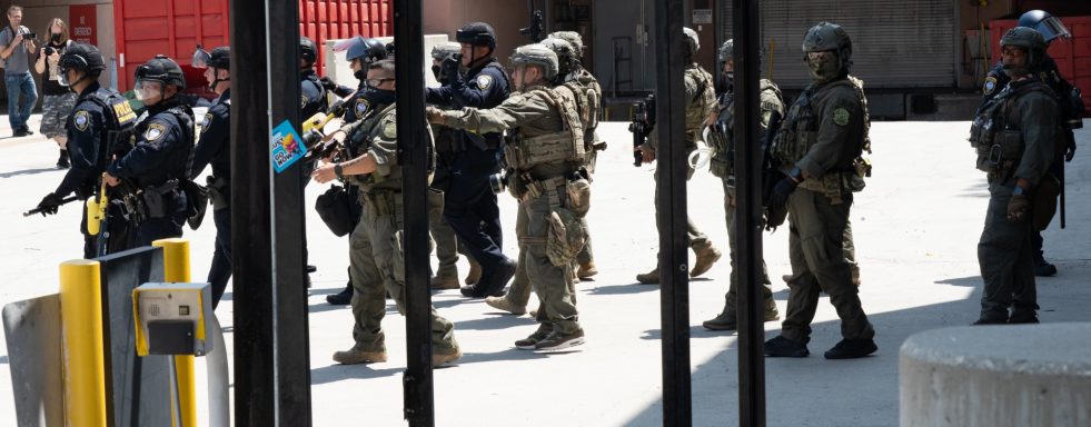 A group of armed police officers in tactical gear at a scene with vehicles.