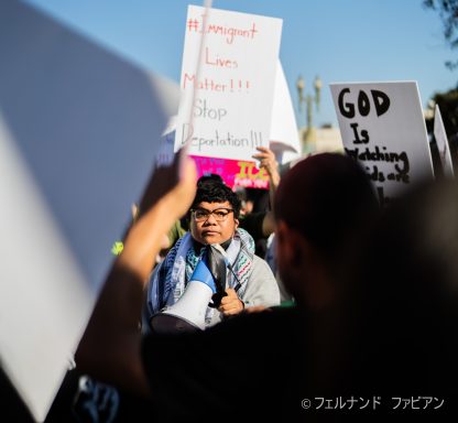 Person with a megaphone at a protest, holding a sign, surrounded by a crowd.