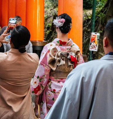 A woman in a pink kimono with floral patterns stands facing a torii gate, surrounded by others.