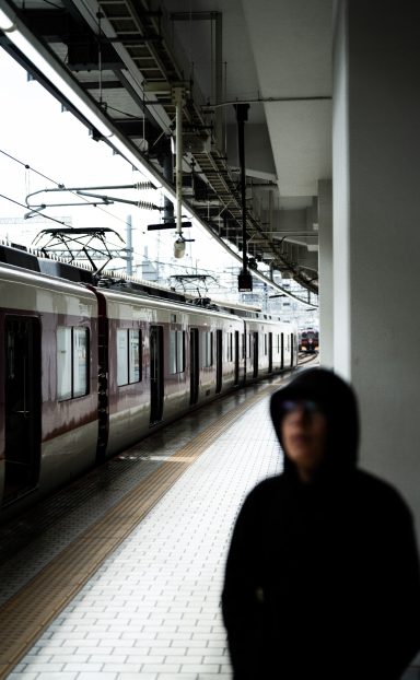 A person in a hooded jacket stands near a train platform, with trains in the background.