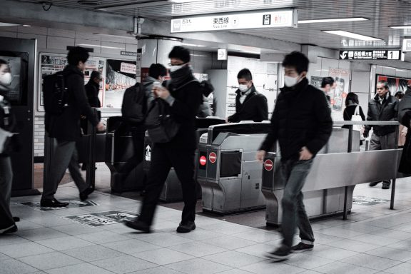 Crowded subway station with commuters wearing masks and passing through turnstiles.