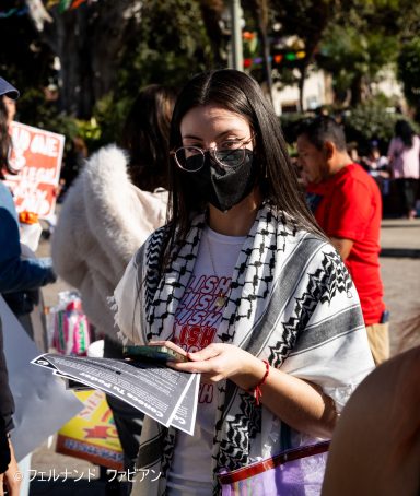 Young woman in a black mask holding a smartphone and a flyer, surrounded by a crowd.